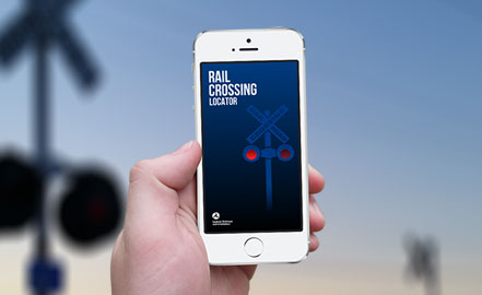 Close up ng smartphone na nagpapakita ng Rail Crossing Locator. Ang background ay isang malabong larawan ng isang tawiran ng riles.