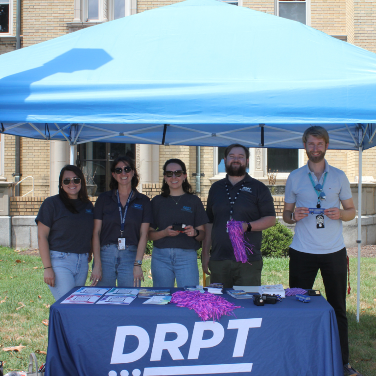 Mga kawani ng DRPT na may hawak na mga pampromosyong bagay sa mesa na may tatak ng DRPT sa ilalim ng asul na tolda