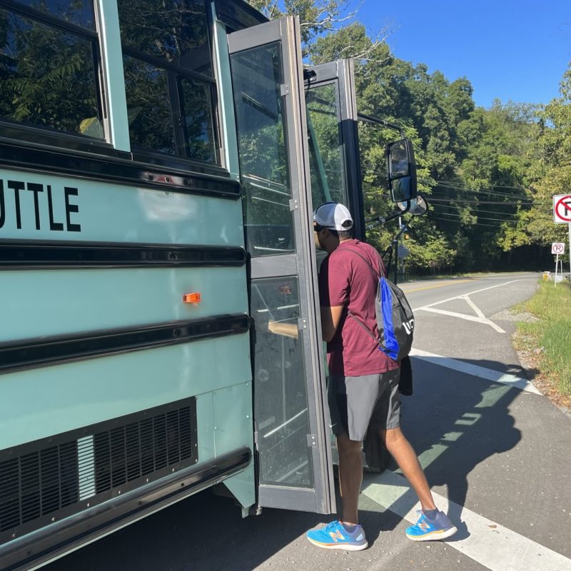 Larawan ng lalaking nakasakay sa bus. Naka-shorts siya, short sleeve shirt, at backpack. Asul na langit at mga puno.